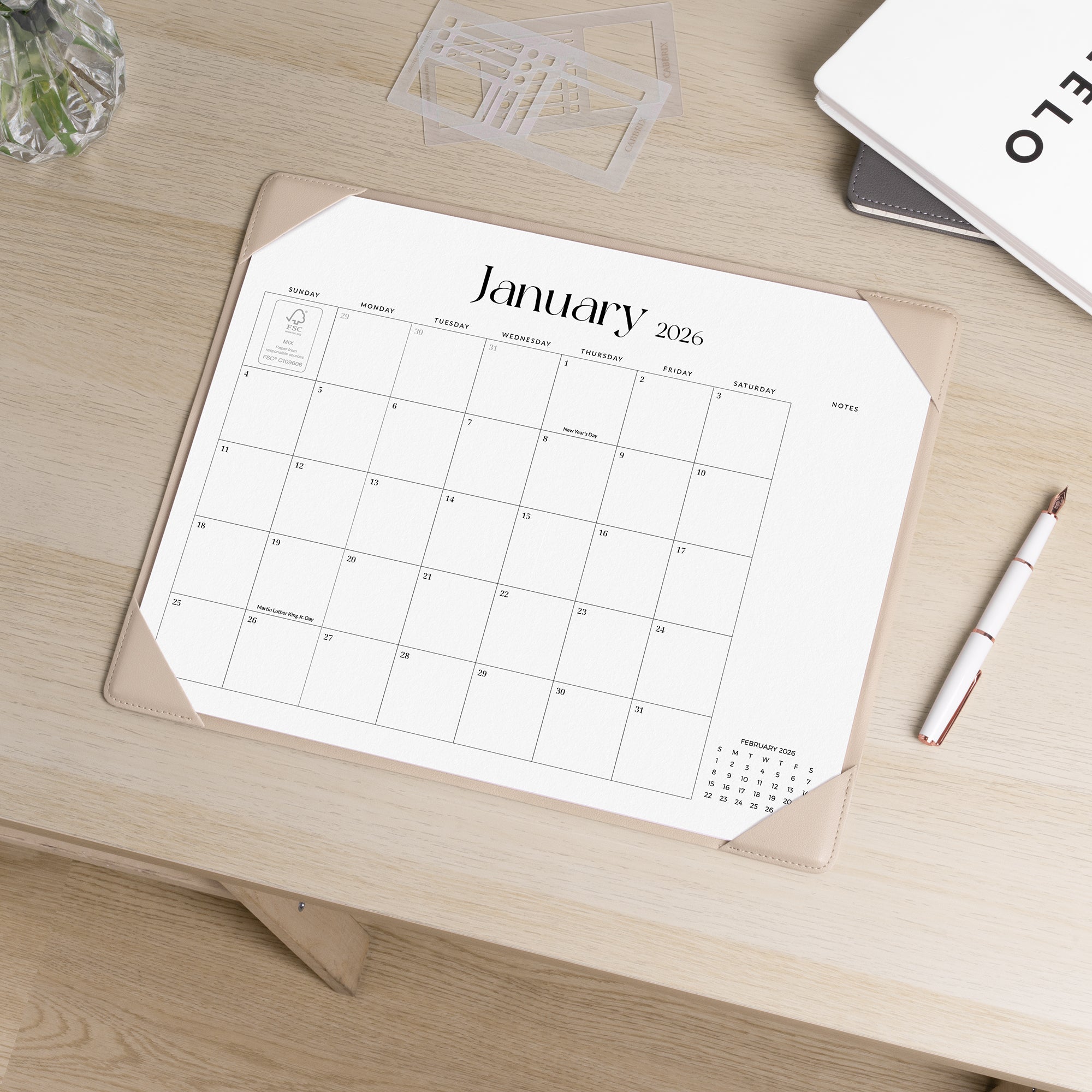 Calendar for January 2026 on a wooden desk with a pen and glass.