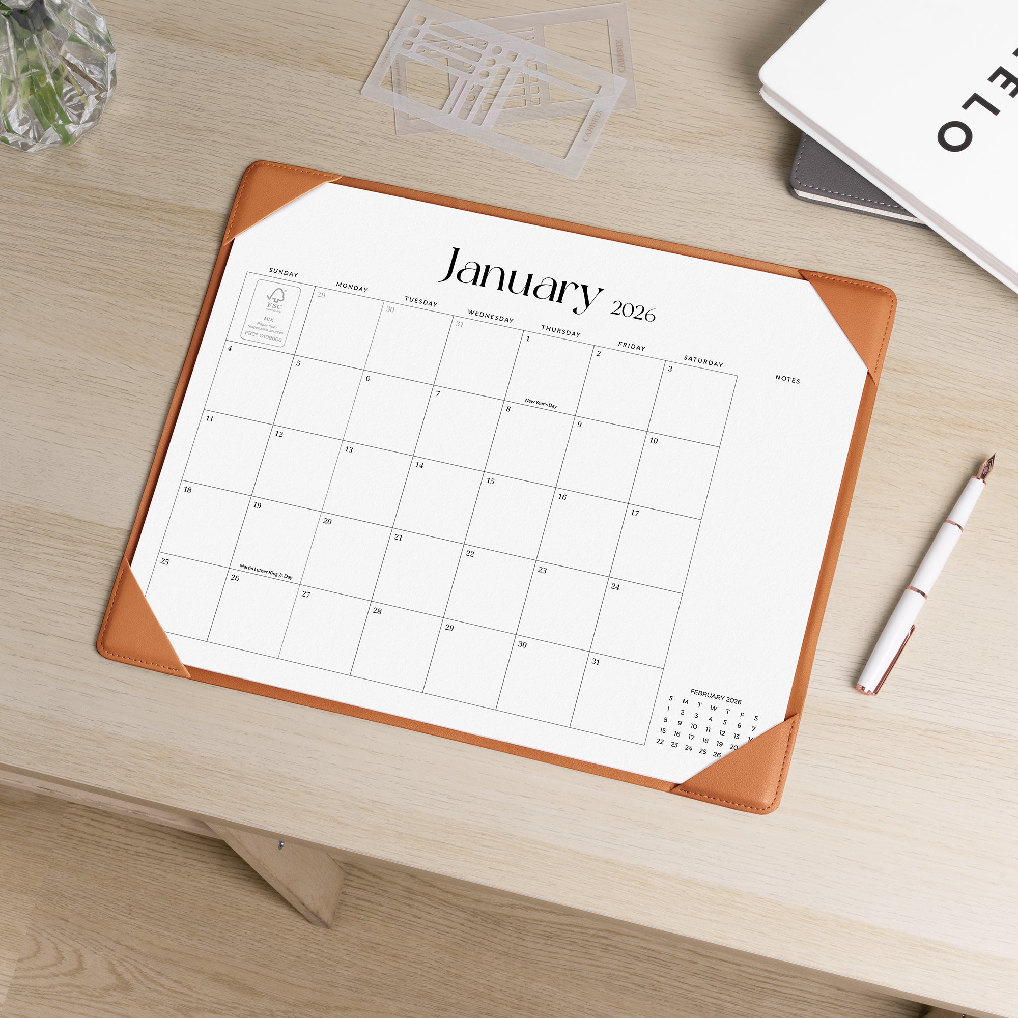 Calendar pad for January 2026 on a wooden desk with a pen and glassware.