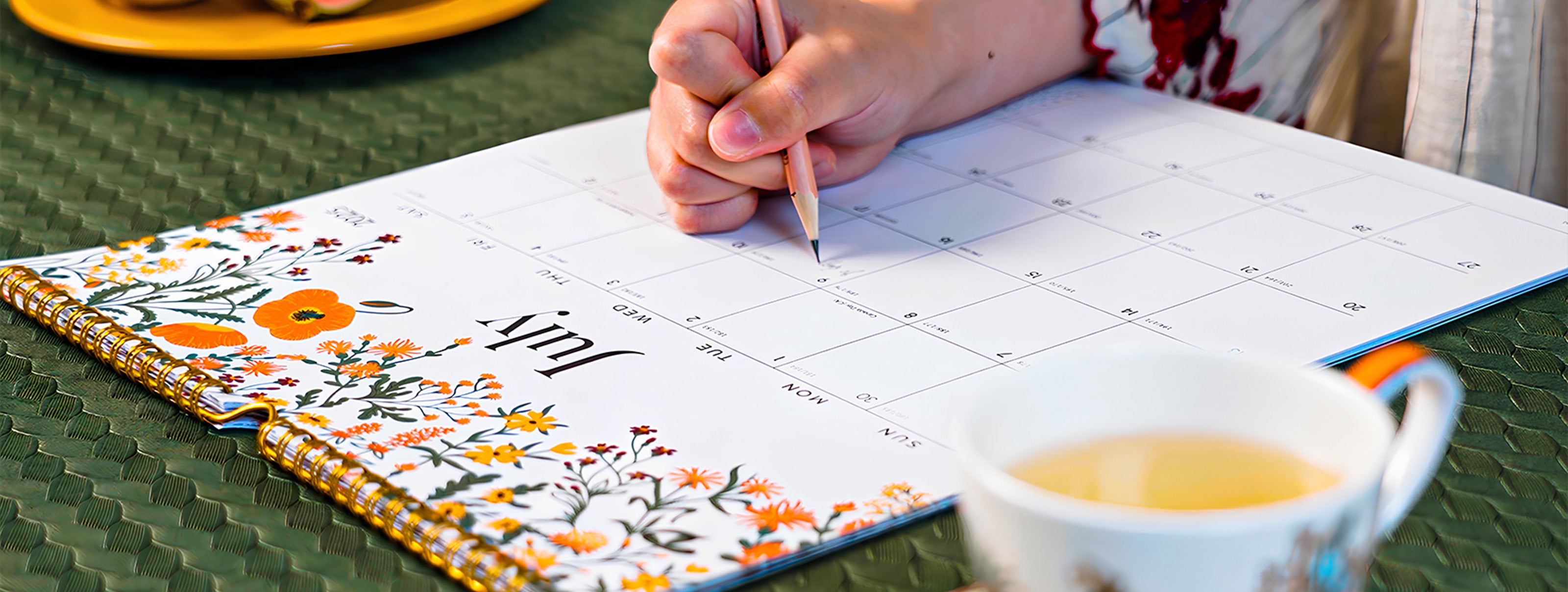 Person writing in a floral notebook with a cup of tea on a green tablecloth